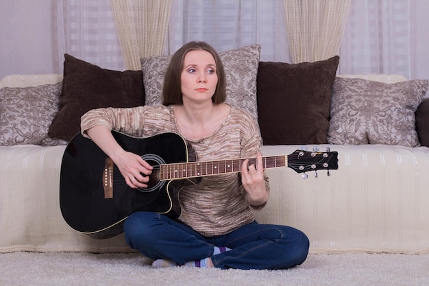 Een jonge vrouw speelt een zwarte akoestische gitaar op het tapijt in de kamer