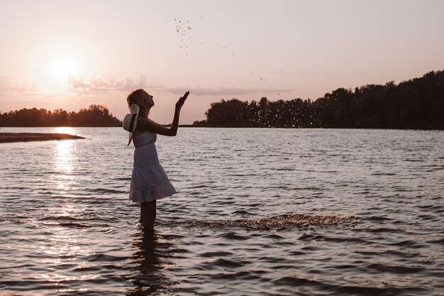 Een jonge vrouw spettert water. Foto van een slanke, mooie, gelukkige blonde in een zomerjurk en een strohoed die in de rivier tegen de zonsondergang staat en haar handen opsteekt.