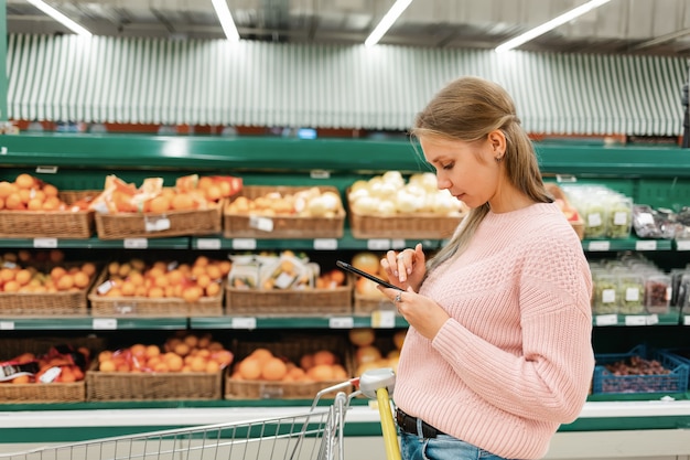 Een jonge vrouw staat in een kruidenierswinkel en leest berichten op haar mobiele telefoon.