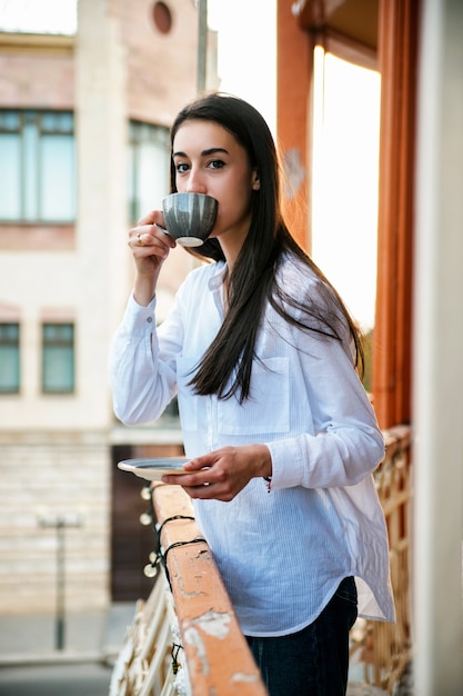 Een jonge vrouw staat op het balkon en drinkt koffie of thee. het meisje is kalm en ontspannen.