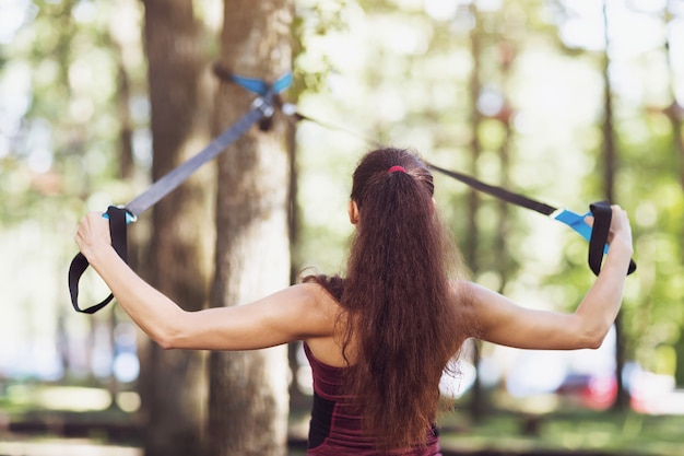 Een jonge vrouw voert een oefening uit om de rugspieren te trainen op een machine boven het hoofd die aan een boom in het park is bevestigd