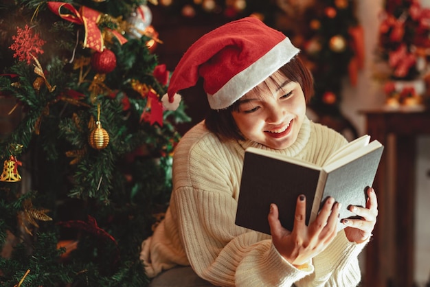 Een jonge vrouw voor de kerstboom die thuis een boek leest met kerstversiering.