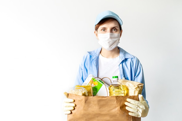 Een jonge vrouw vrijwilliger in een blauw uniform en een medisch masker en handschoenen houdt een papieren zak met voedsel.