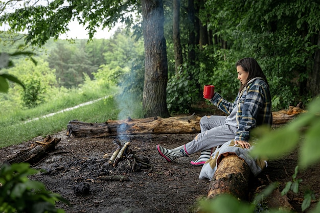 Een jonge vrouw warmt zich op bij een uitgedoofd vuur met een kopje verwarmende drank in het bos tussen de bomen.