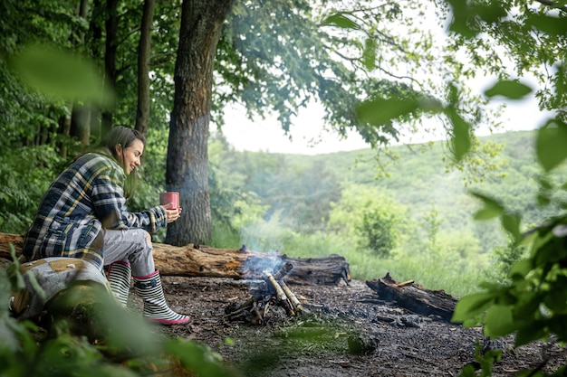 Een jonge vrouw warmt zich op bij een uitgedoofd vuur met een kopje verwarmende drank in het bos tussen de bomen.