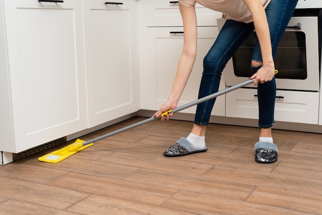 Een jonge vrouw wast houten vloeren van een laminaat in een lichte keuken. Het meisje wast stof en vuil onder de keukenkast vandaan