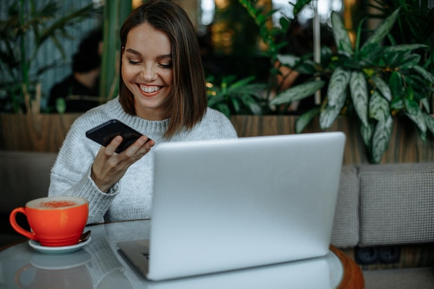 Een jonge vrouw werkt aan een laptop in een café en neemt een audioboodschap op op een smartphone