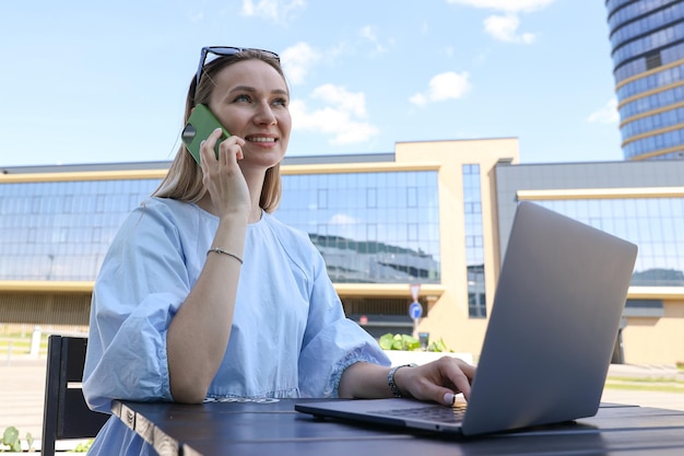 Een jonge vrouw werkt op een laptop en telefoneert op een zomerterras