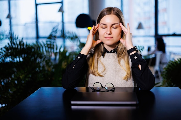 Foto een jonge vrouw zit aan een tafel met een gesloten laptop en masseert haar hoofd van hard werken en hoofdpijn vrouw die lijdt aan migraine