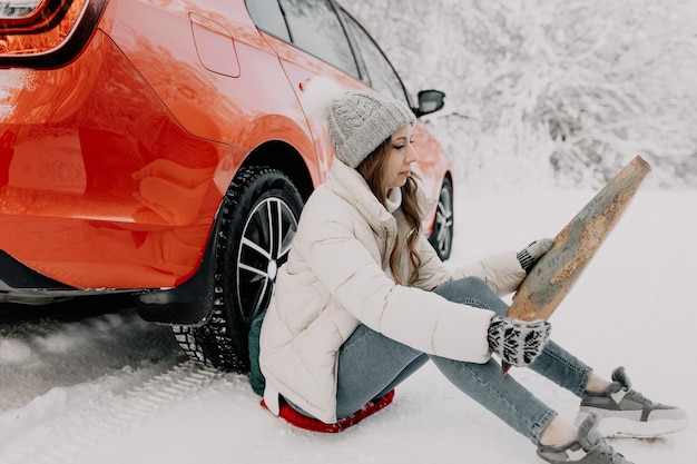 Een jonge vrouw zit bij een rode auto en heeft een kaart in haar handen. Reis concept.