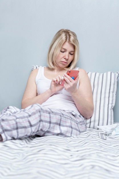 Een jonge vrouw zit in bed met een thermometer en een telefoon Onaangename symptomen van de ziekte en een doktersconsult Blonde in pyjama tegen een grijze muur Verticaal