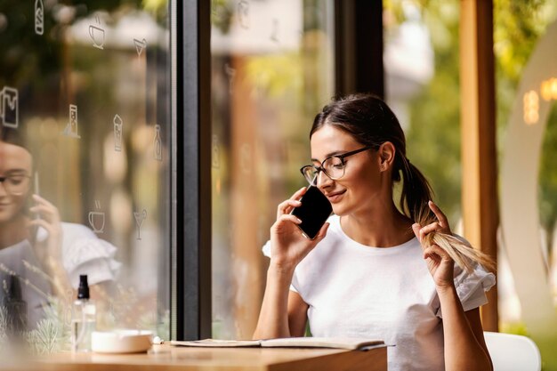 Een jonge vrouw zit in een cafetaria en kijkt naar het menu en praat aan de telefoon