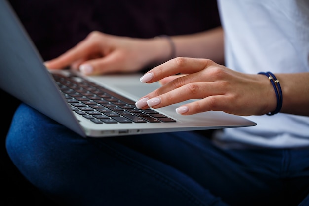 Een jonge vrouw zit op de bank en werkt op afstand van haar werk op een laptop. Meisje met een computer op haar knieën die naar de monitor kijkt