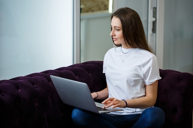 Een jonge vrouw zit op de bank en werkt op afstand van haar werk op een laptop. Meisje met een computer op haar knieën die naar de monitor kijkt