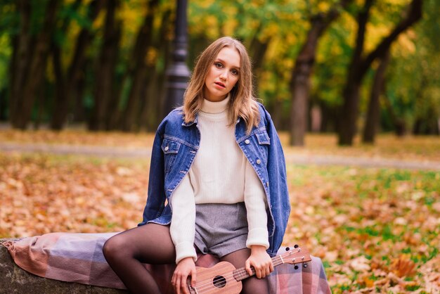 Een jonge vrouw zit op de bank in het park en speelt ukelele, poseren met gitaar