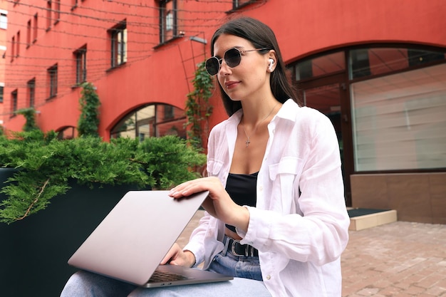 Een jonge vrouw zit op een bankje en werkt met een laptop op straat