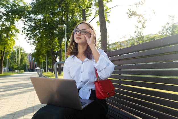 Een jonge vrouw zit op een bankje en werkt op een laptop in het park