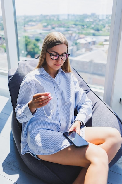 Een jonge vrouw zit op een fauteuil en luistert naar muziek op een koptelefoon met behulp van een telefoon Mooie vrouw ontspant na een werkdag ontspannen in een coworking-ruimte met grote ramen