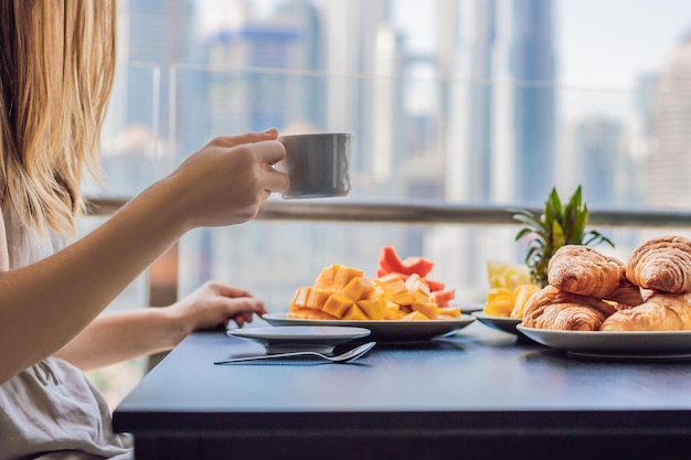 Een jonge vrouw zit op het balkon te ontbijten. Ontbijttafel met koffiefruit en broodcroisant op een balkon tegen de achtergrond van de grote stad