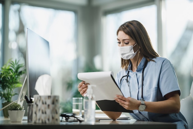 Een jonge vrouwelijke arts met beschermend gezichtsmasker die alleen zit en in haar spreekkamer werkt tijdens een pandemie van het coronavirus.