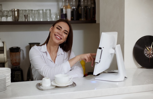 Een jonge vrouwelijke barista drukt de bestelling achter de bar af en kijkt glimlachend