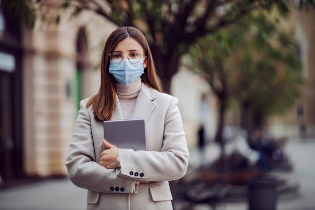 Een jonge vrouwelijke freelancer met gezichtsmasker op straat staan en tablet vasthouden