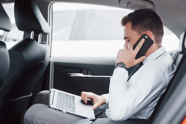 Een jonge zakenman die aan laptop werkt en op de telefoon spreekt terwijl het zitten in de rug van de auto. Werkt in beweging, waardeert zijn tijd