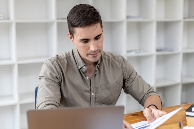 Een jonge zakenman die op kantoor zit en met een laptop werkt, hij controleert financiële documenten, hij heeft een startup opgericht, hij is een jonge ondernemer die de vaardigheden heeft om een groeiend bedrijf te leiden.