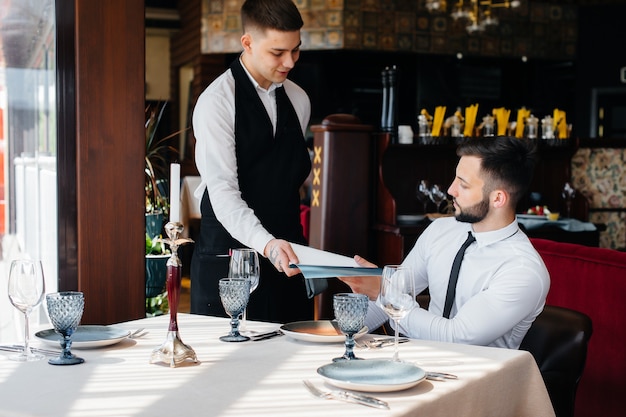 Een jonge zakenman in een goed restaurant bekijkt het menu en bestelt een jonge ober in een stijlvol schort. Klantenservice.