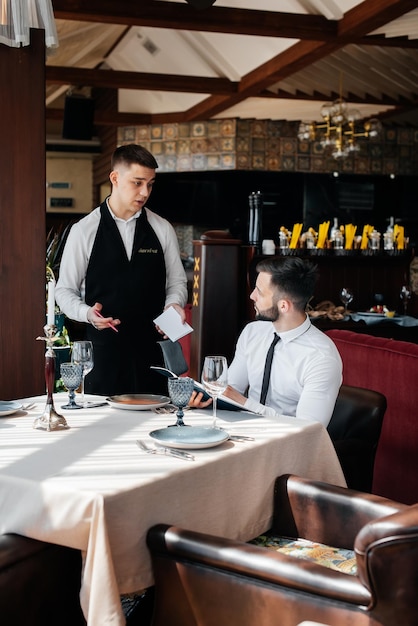 Een jonge zakenman in een goed restaurant onderzoekt het menu en plaatst een bestelling bij een jonge ober in een stijlvol schort klantenservice service aan tafel in het restaurant