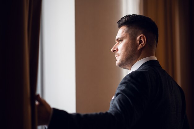 Een jonge zakenman in een pak die de gordijnen in zijn hotelkamer scheidt om het ochtendlicht binnen te laten