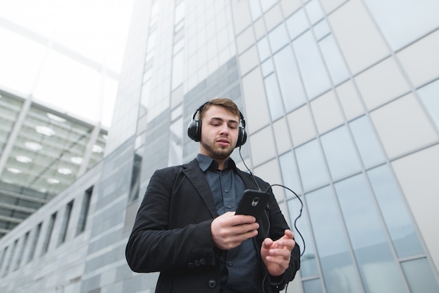 Een jonge zakenman luistert naar muziek in een koptelefoon en gebruikt een smartphone terwijl hij door de stad loopt.