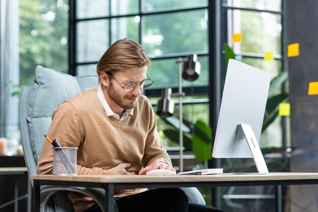 Een jonge zakenman programmeur kantoormedewerker zit aan tafel en houdt zijn maag vast, hij voelt zich ernstig