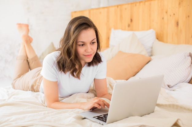 Een jonge zakenvrouw kijkt naar het scherm van een laptop die op internet werkt, het meisje ligt op het bed en gebruikt een laptop