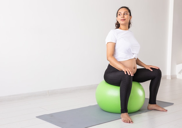 Een jonge zwangere vrouw die ontspanningsoefeningen doet met een fitnessbal terwijl ze op een mat zit