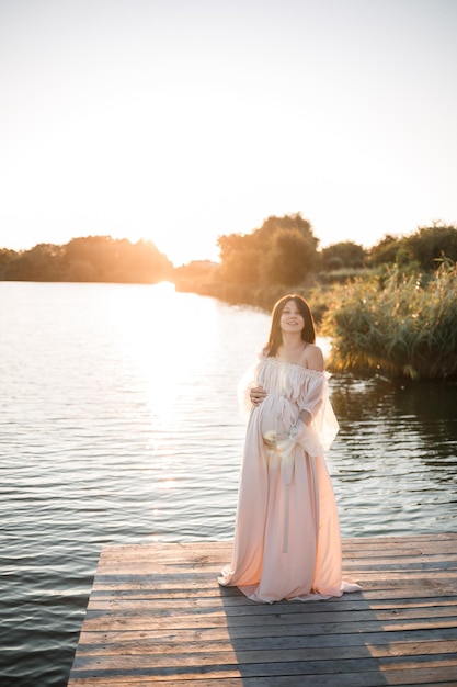 Een jonge zwangere vrouw in een jurk van chiffon staat op een pier bij de rivier tegen de achtergrond van een oranje zonsondergang