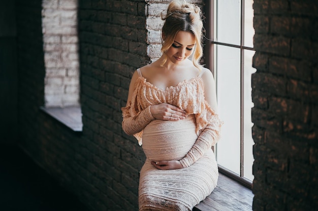 Een jonge zwangere vrouw in een mooie jurk zit voor het raam
