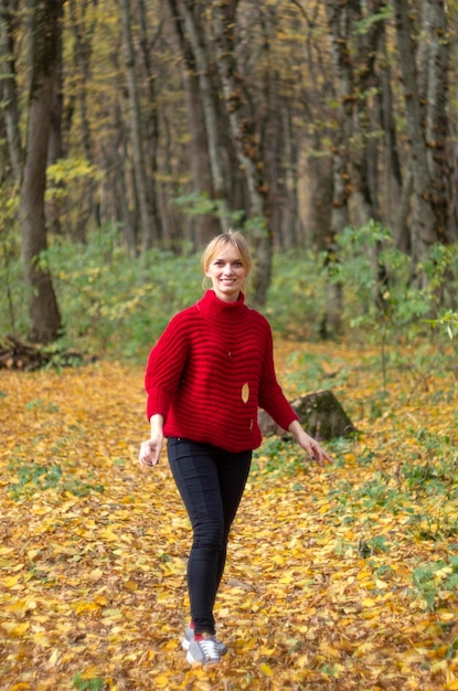Een jonge zwangere vrouw in een rode trui tussen gevallen bladeren in een herfstbos in een goed humeur