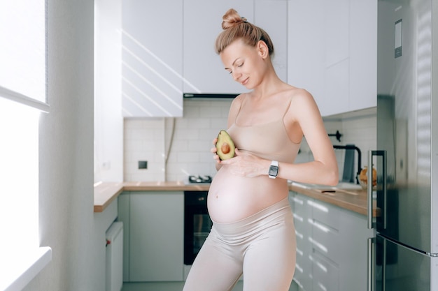 Een jonge zwangere vrouw van Europees uiterlijk in haar keuken met een avocado in haar handen het concept van goede voeding van zwangere vrouwen voor de juiste ontwikkeling van de foetus