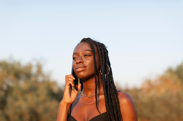 Een jonge zwarte vrouw met vlechten in haar haar praat gelukkig op haar smartphone tijdens een wandeling bij zonsondergang