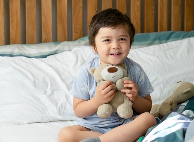 Een jongen die met het glimlachen gezicht in de ochtend ontwaakt, Gelukkig kind dat met teddybeer i speelt