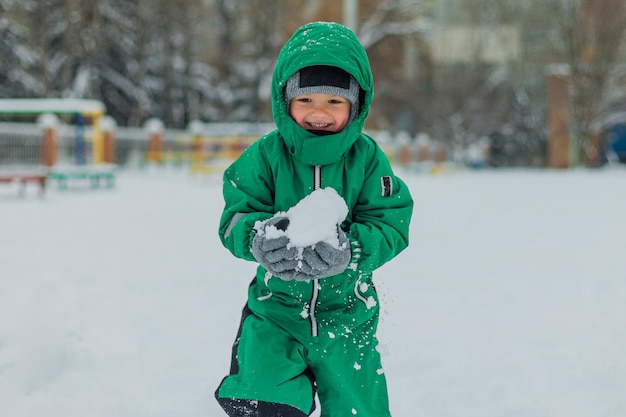 Foto een jongen draagt sneeuw in zijn handen jongens groene jumpsuit draagt sneeuw kleine jongen die in de sneeuw loopt