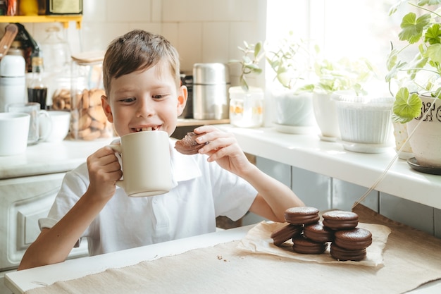 Een jongen drinkt melk en chocoladekoekjes in zijn thuiskeuken. Gelukkig kind drinkt melk uit een witte kop en eet schooltaarten.