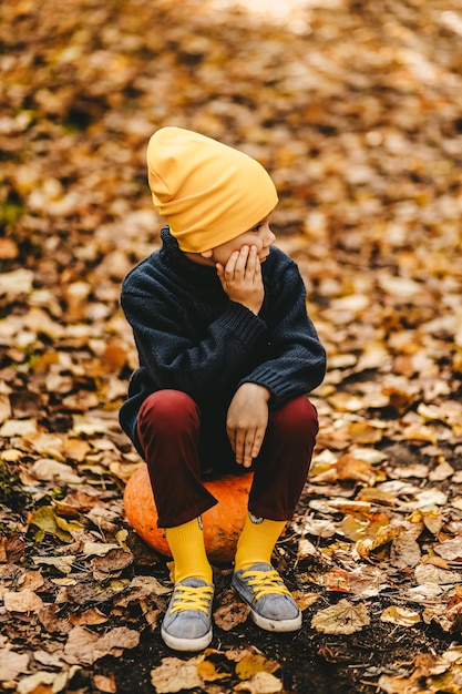 Foto een jongen een tienerkind in een heldere gele hoed zit op een pompoen in een herfstbos in de natuur buiten