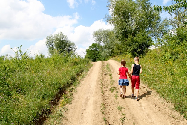 Een jongen en een meisje kinderen lopen op een onverharde weg op een zonnige zomerdag. kinderen houden handen samen terwijl u geniet van ativity buitenshuis.