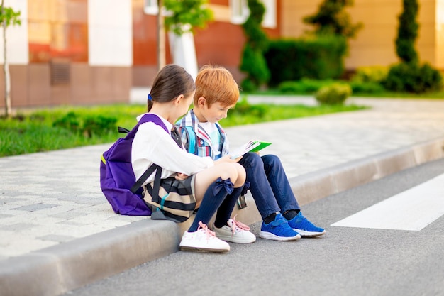 Een jongen en een meisje met aktetassen of rugzakken zitten op school en lezen een boek en tablet die terug naar school gaan broer en zus studeren huiswerk op straat