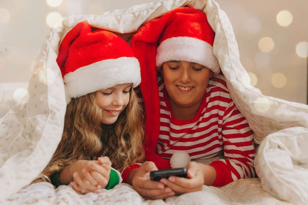 Een jongen en een meisje met kerstmutsen liggen onder de dekens met smartphones in hun handen. Nieuwjaar en Kerstmis