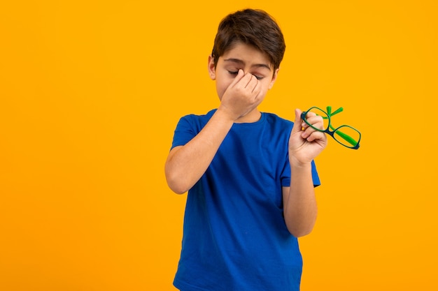 Een jongen in een blauw T-shirt houdt een bril in zijn hand en wrijft zijn ogen op geel met kopie ruimte