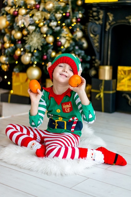 Een jongen in een elfkostuum met mandarijnen bij de kerstboom wacht op Kerstmis