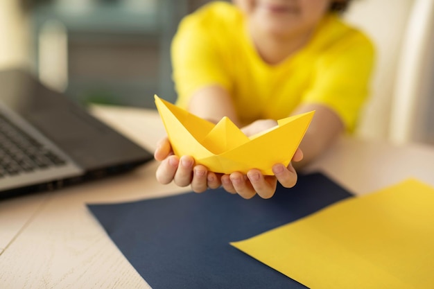 een jongen in een geel T-shirt bij een online origami-workshop een papieren bootje